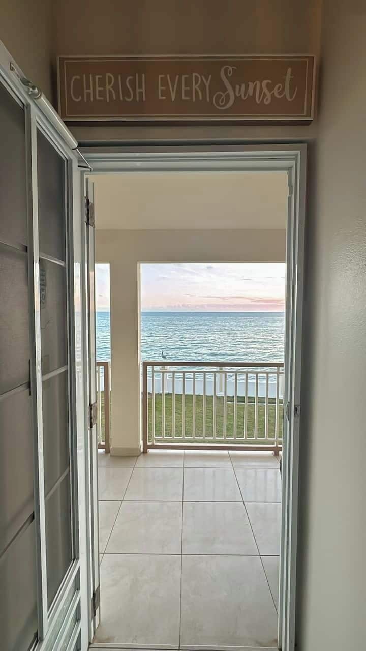 Eine offene Tür führt zu einem Balkon mit Blick auf das Meer, darüber ein Schild mit der Aufschrift 'CHERISH EVERY Sunset'.