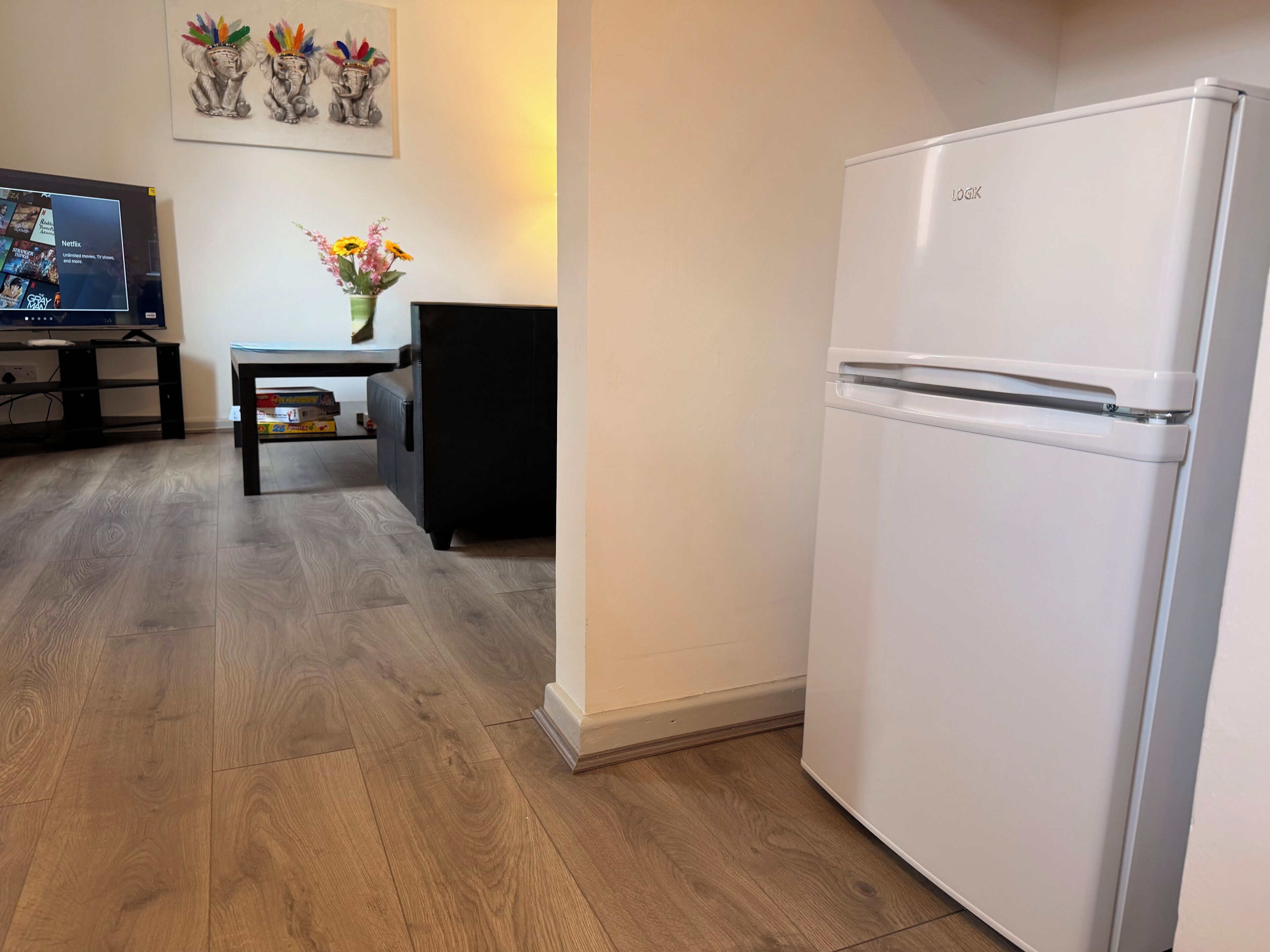 A modern kitchen with a white refrigerator, a wooden dining table with a vase of flowers, and a flat-screen TV on a stand.