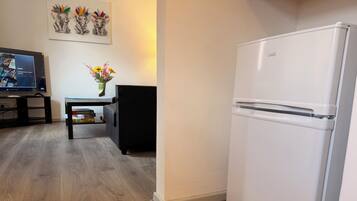 A modern kitchen with a white refrigerator, a wooden dining table with a vase of flowers, and a flat-screen TV on a stand.