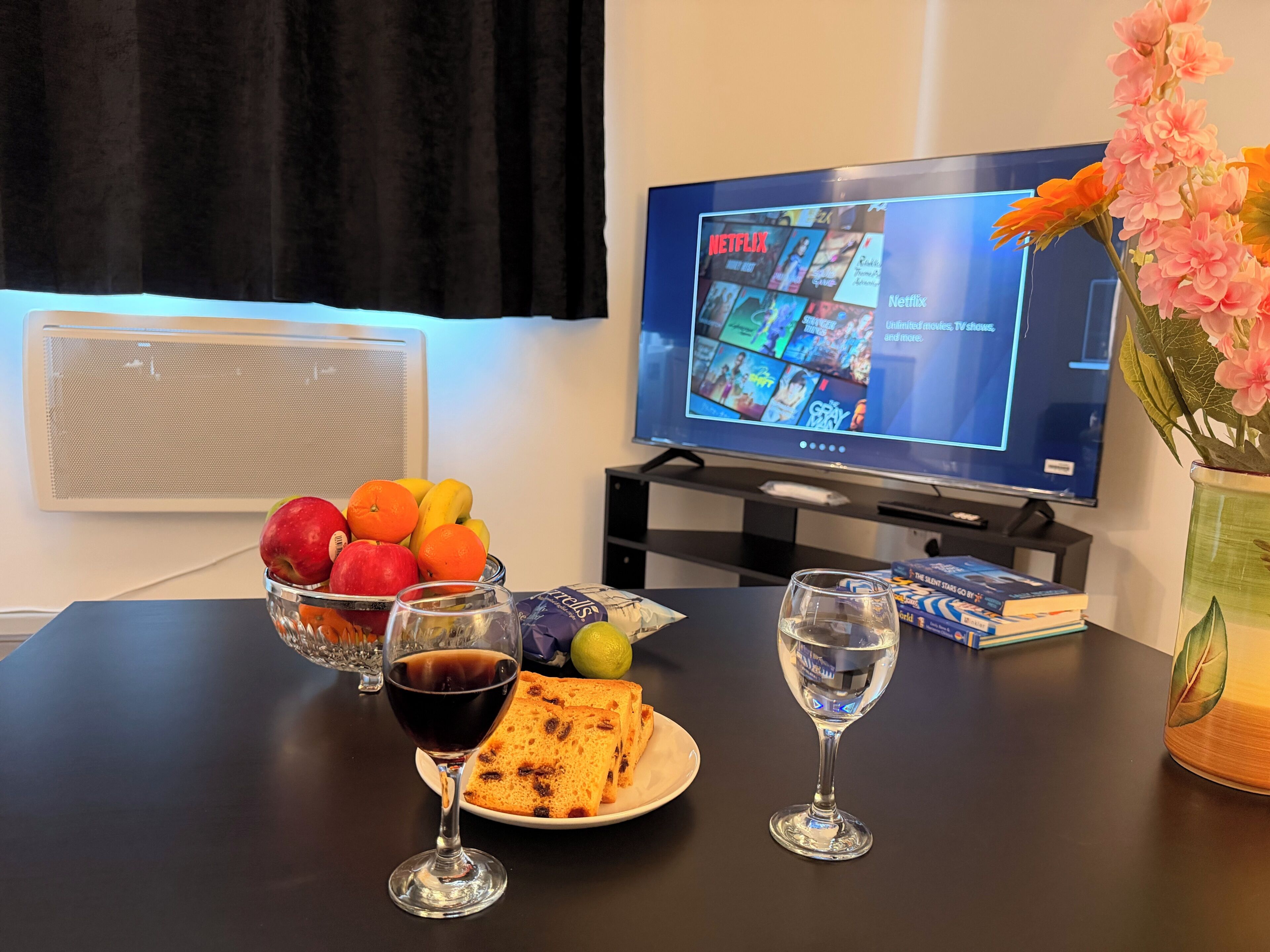 Une table avec des fruits, un verre de vin rouge, une assiette de toasts, un verre d’eau, un vase de fleurs et un téléviseur diffusant Netflix.