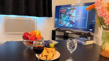 Une table avec des fruits, un verre de vin rouge, une assiette de toasts, un verre d’eau, un vase de fleurs et un téléviseur diffusant Netflix.