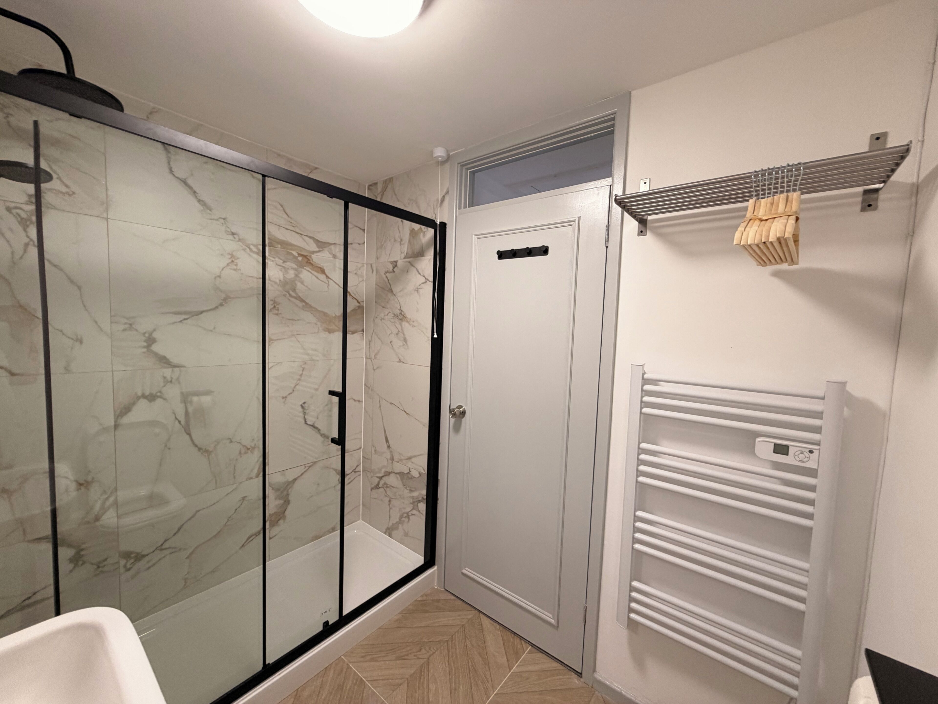 Baño con cabina de ducha de vidrio, puerta blanca y toallero con termómetro digital.