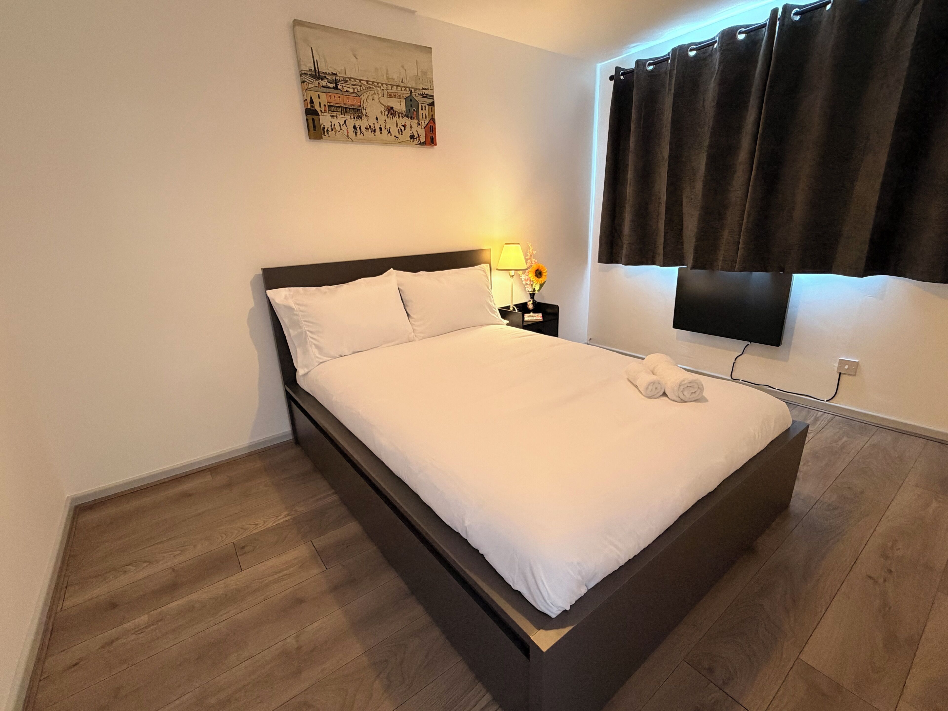 A neatly made bed in a hotel room with a flat-screen TV mounted on the wall, a painting above the bed, and a dark curtain on the window.