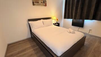 A neatly made bed in a hotel room with a flat-screen TV mounted on the wall, a painting above the bed, and a dark curtain on the window.