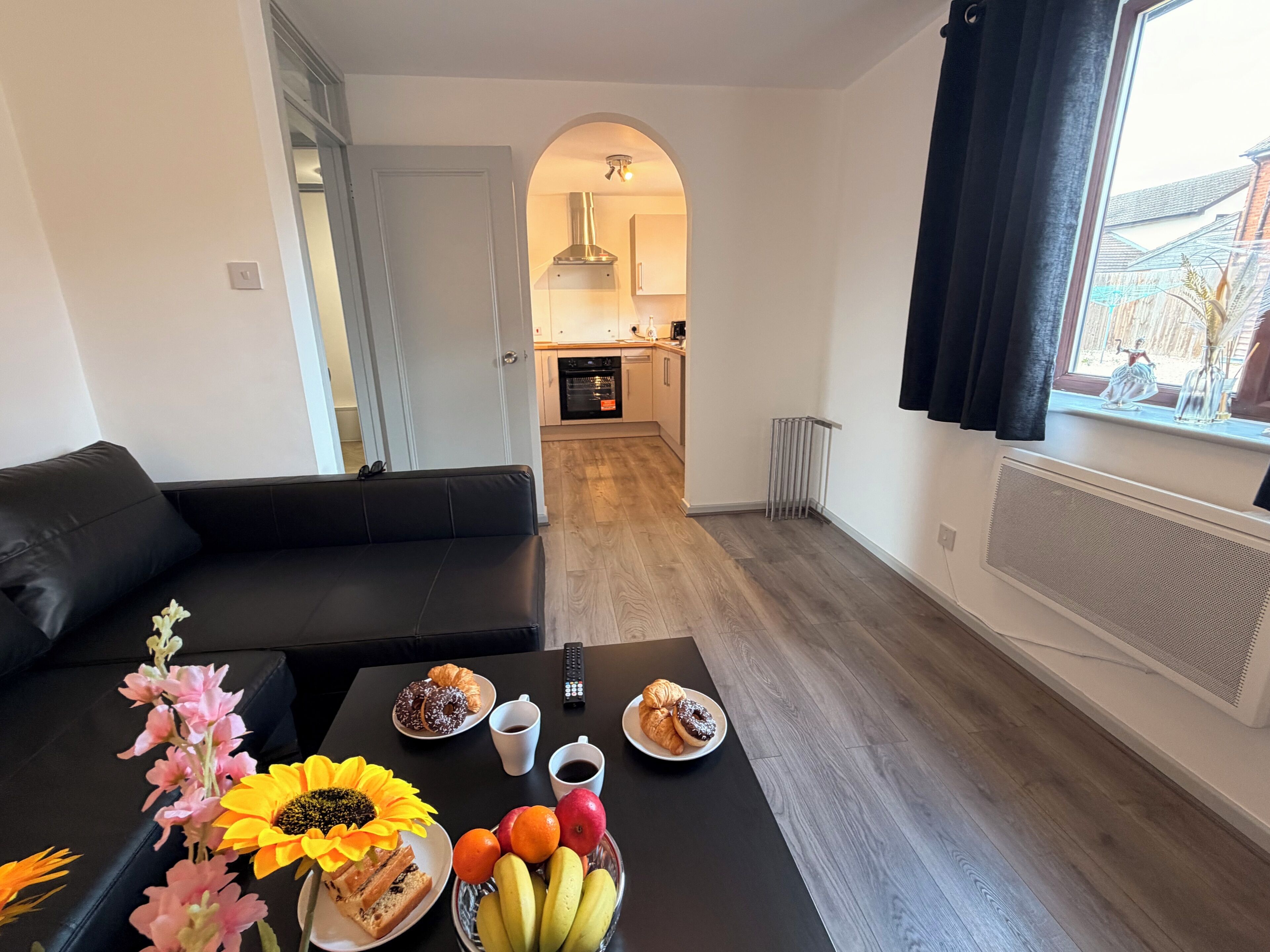 Una sala de estar moderna con un sofá negro, una mesa de comedor con comida y bebidas, y vista hacia la cocina.
