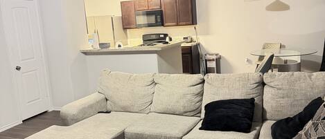 A living room with a sectional sofa, a white coffee table, and a kitchen in the background.