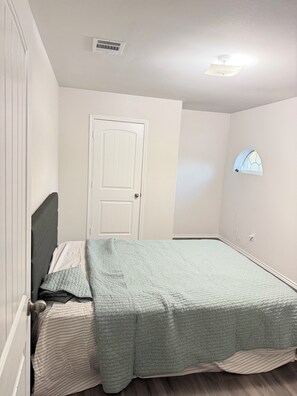 Un dormitorio con cama, puerta, ventana y rejilla de ventilación en el techo.