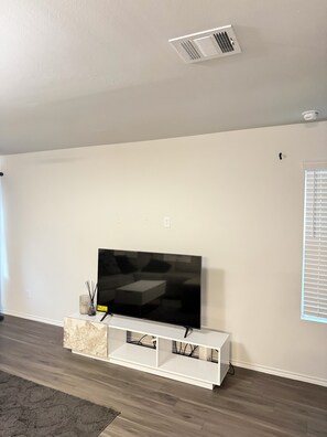 A living room with a flat-screen TV on a white stand, a ceiling vent, and a window with blinds. -  A Peaceful Richmond Stay in Houston Made for Comfort & Memories. (Richmond)