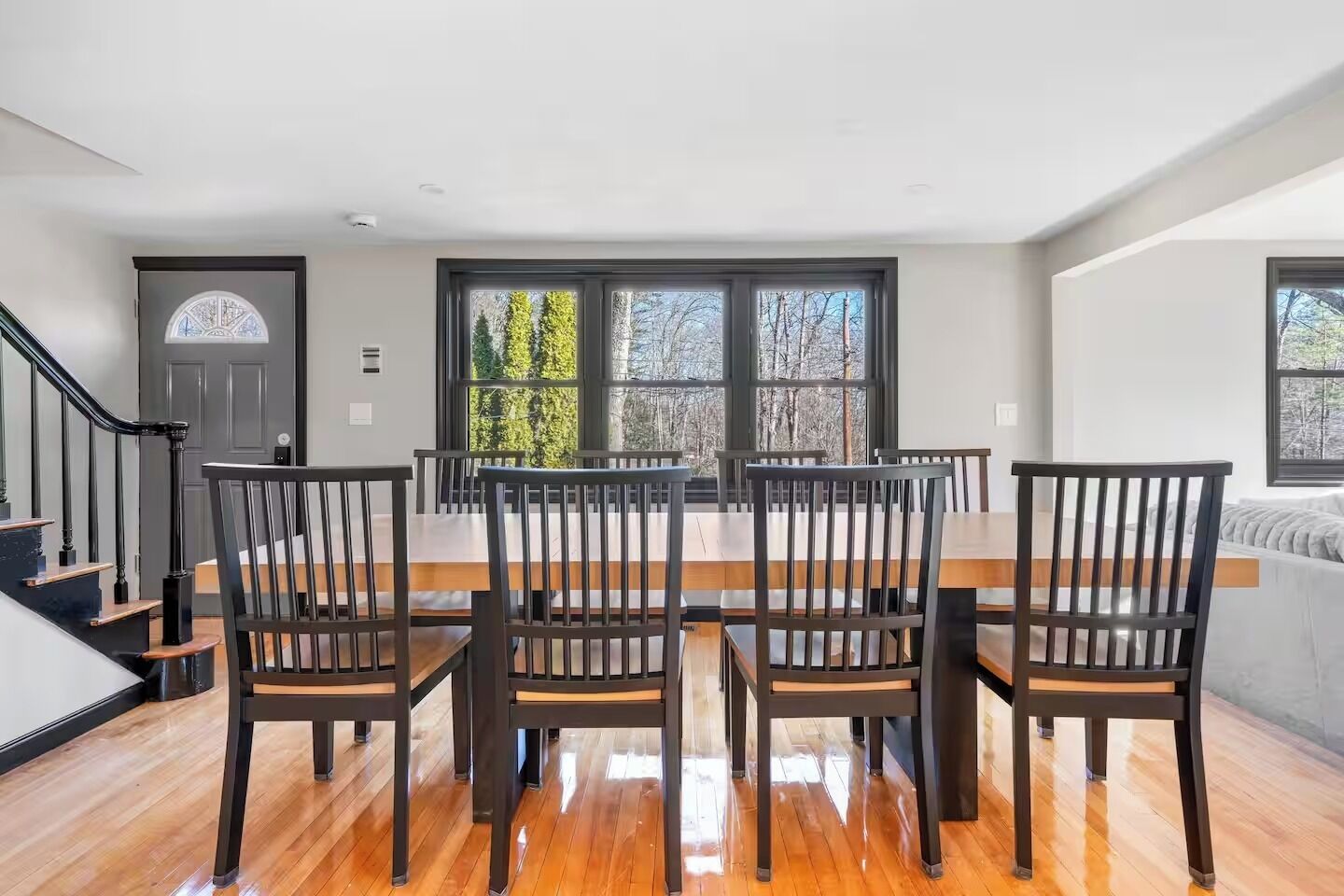 Un comedor con una mesa y sillas de madera, ventanas amplias con vistas a árboles y una escalera.