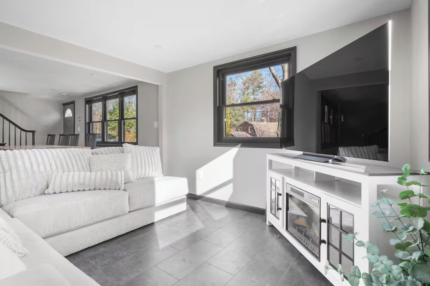 A modern living room with a white sofa, a flat-screen TV, and a large window with a view of the outside.