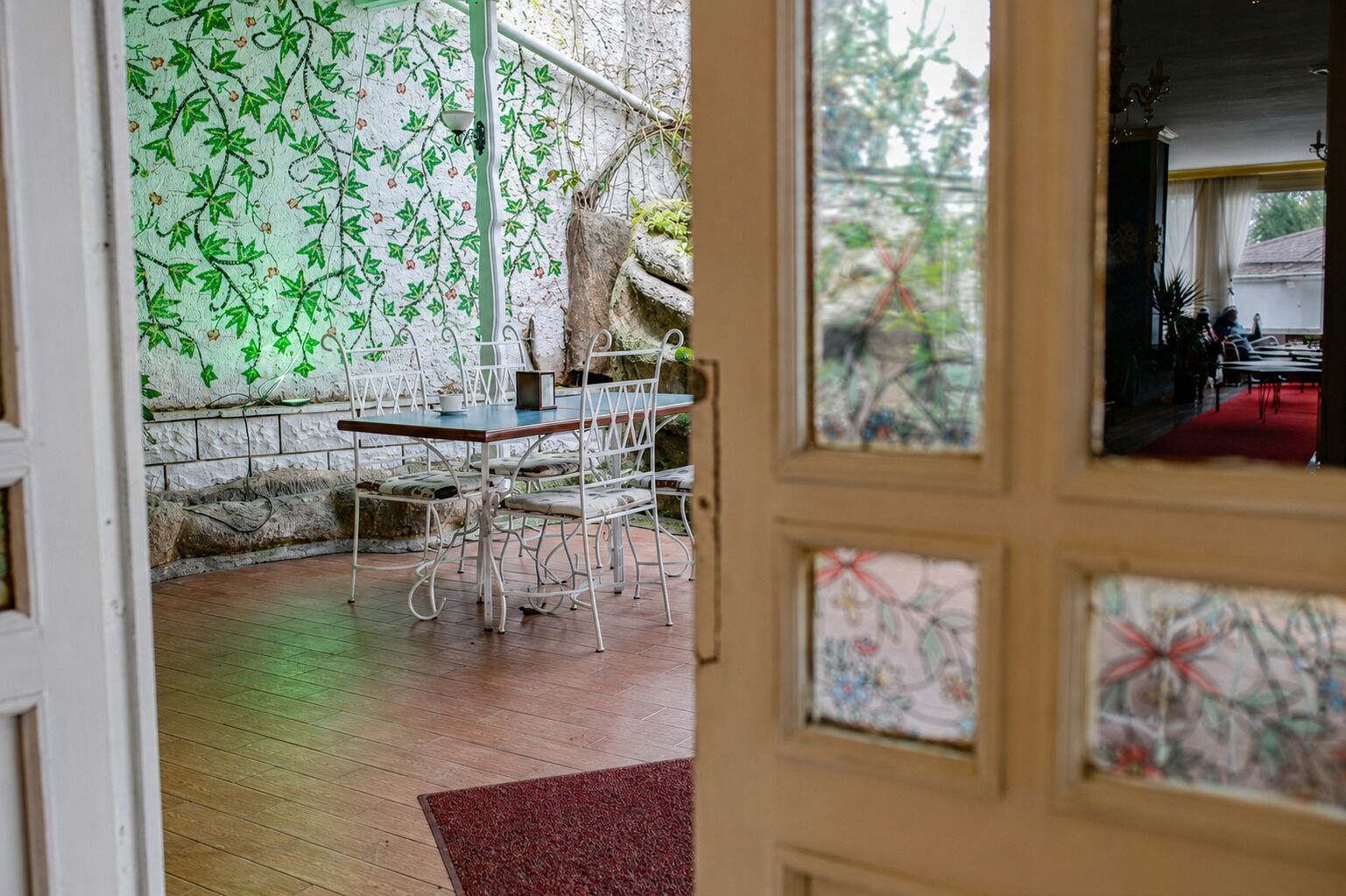 Une pièce comprenant un coin repas avec une table et des chaises, un mur en pierre orné d’un motif végétal, et une vue sur une autre pièce, accessible par une porte.