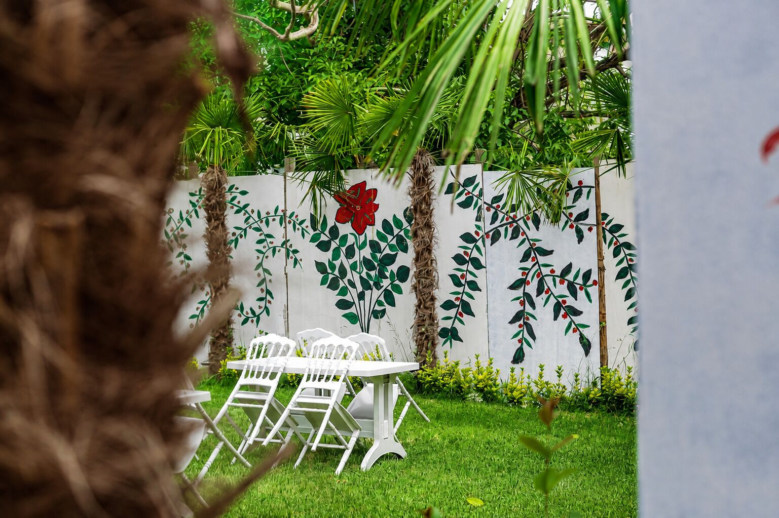 Een tuin met een witte tafel en stoelen, omgeven door weelderige begroeiing en een muur met bladmotieven.