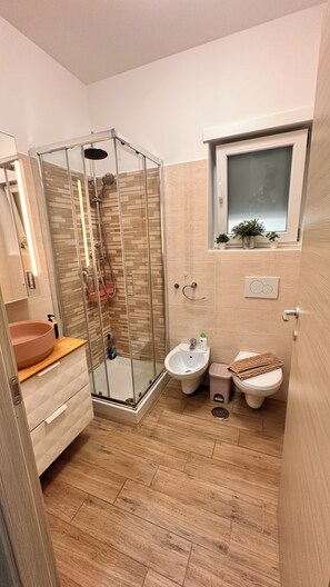 A modern bathroom with a glass-enclosed shower, a vanity with a pink basin, a bidet, and a window above the toilet. - KAIRòS HOME NEAR THE BEACH, PARKING SPACE, BICYCLES, AND BEACH UMBRELLA (Tortoreto lido)