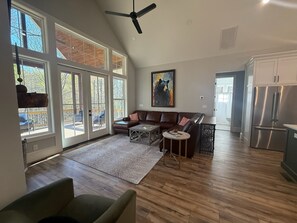 Amplia sala de estar con un gran ventanal, ventilador de techo, sofá, mesa de centro y cocina con refrigerador.