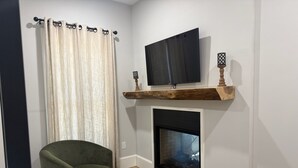 Una sala de estar con un sillón verde, una mesa de centro de vidrio, una chimenea y un televisor montado.