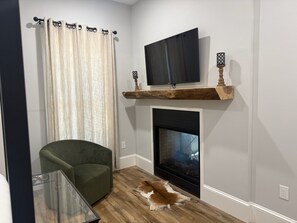 Ein Wohnzimmer mit einem grünen Sessel, einem Glastisch, einem Kamin und einem an der Wand montierten Fernseher.