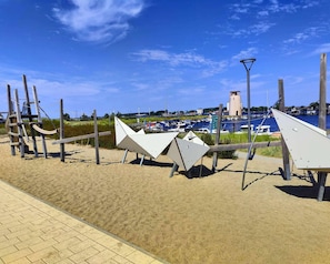 A beach with a sandy walkway, a lighthouse, and boats in the background. - Apartment am Sudstrand in Fehmarn-burgtiefe (Fehmarn)