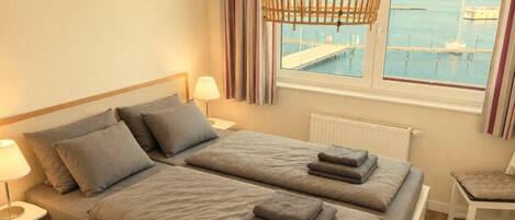 A double bed with a headboard, bedside lamps, a view of a marina through a window, and a wicker light fixture.