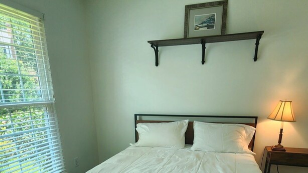 Een slaapkamer met een bed, een raam met zonwering, een nachtkastje met een lamp, en een wandplank met een foto.