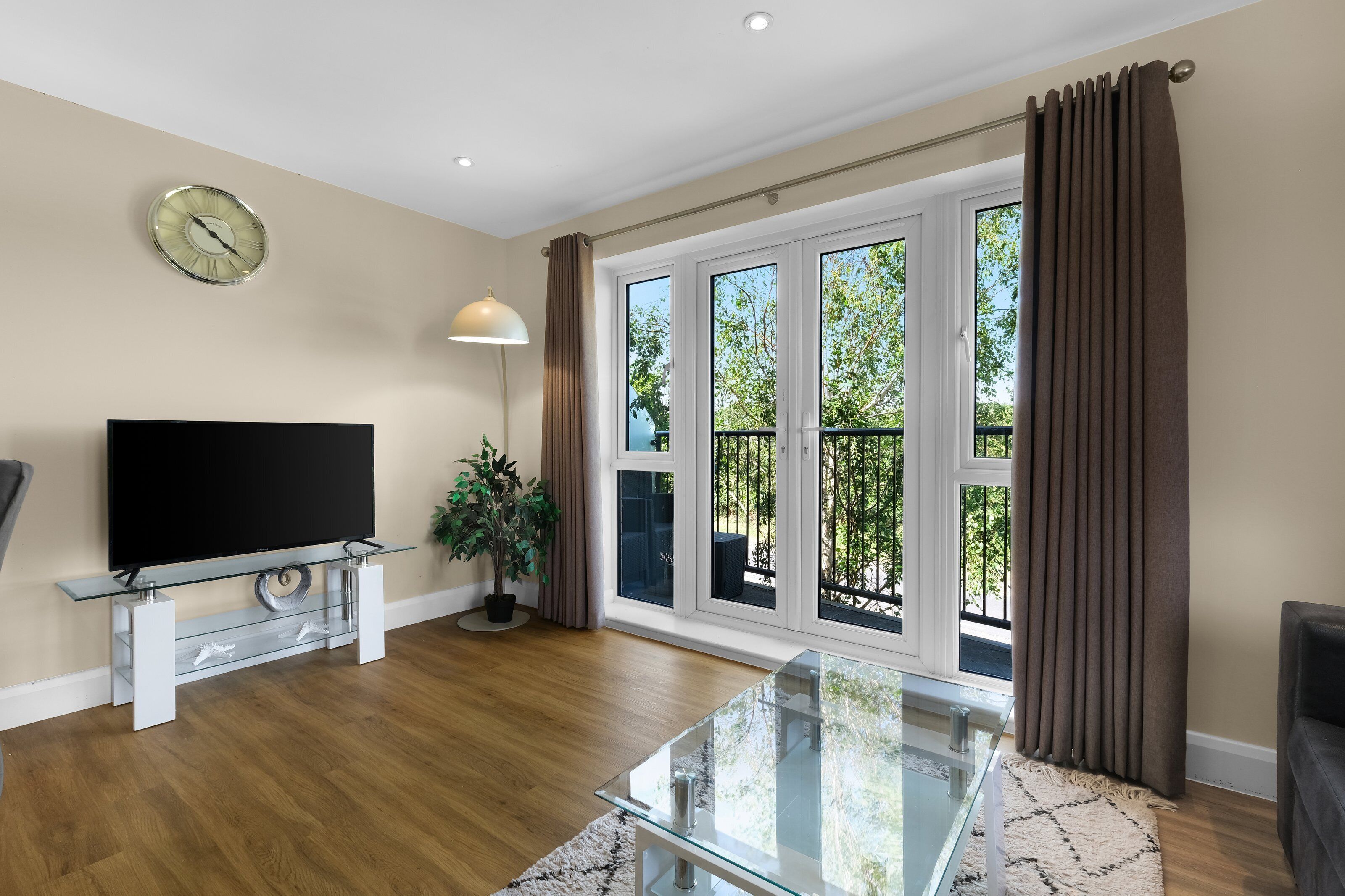 A living room with a flat-screen TV on a glass table, a potted plant, and a large window with curtains.