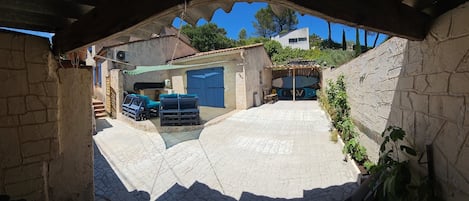 Un patio con suelo de piedra, una zona de estar cubierta y un edificio con puerta azul.