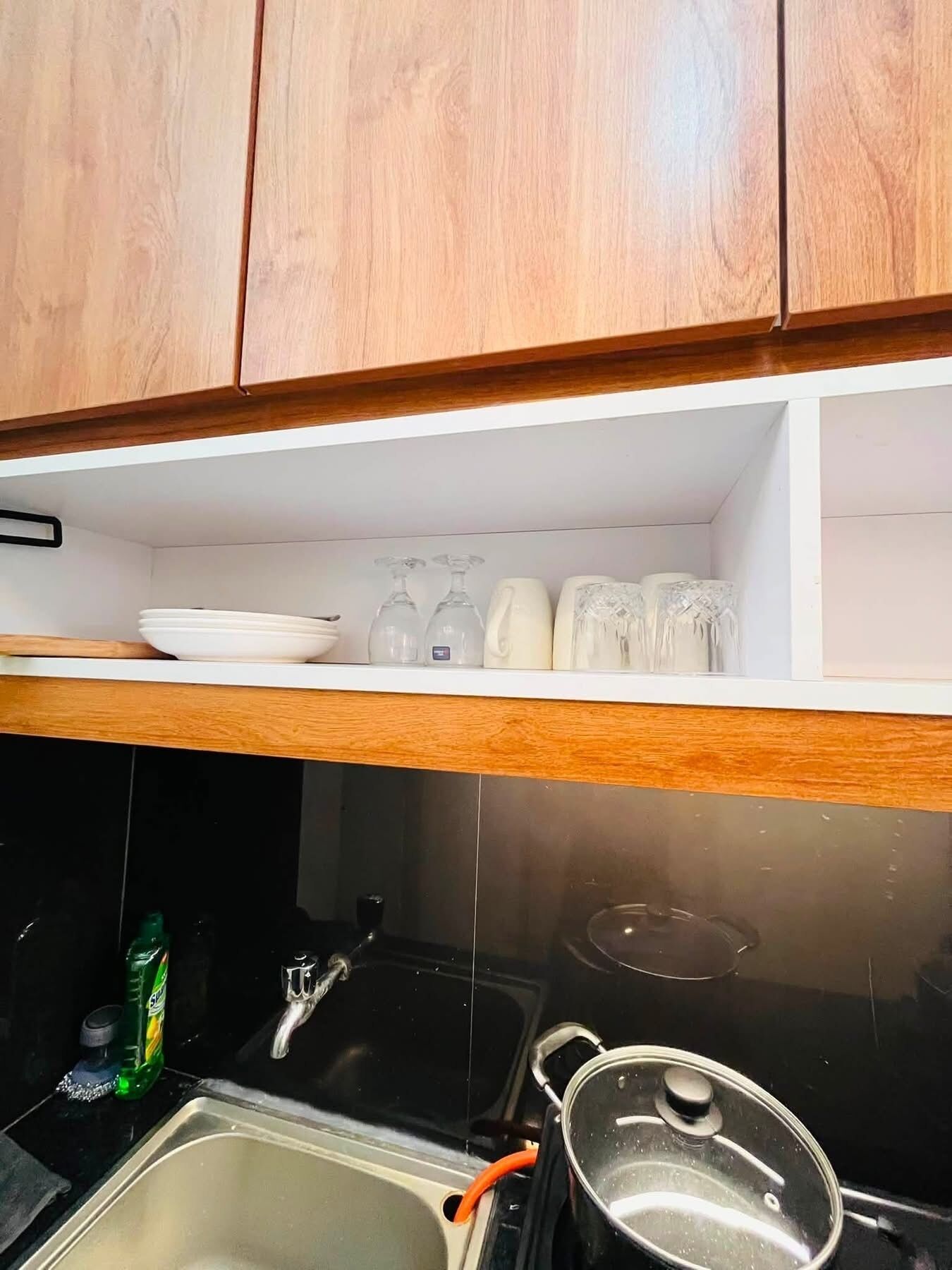A kitchen with wooden cabinets, a stainless steel sink, a pot on the stove, and a shelf with glassware and plates.