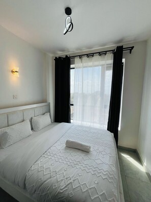 A bedroom with a bed, a wall-mounted light, a window with curtains, and a view outside. - Fabulous 1-bedroom apartment (Nairobi)