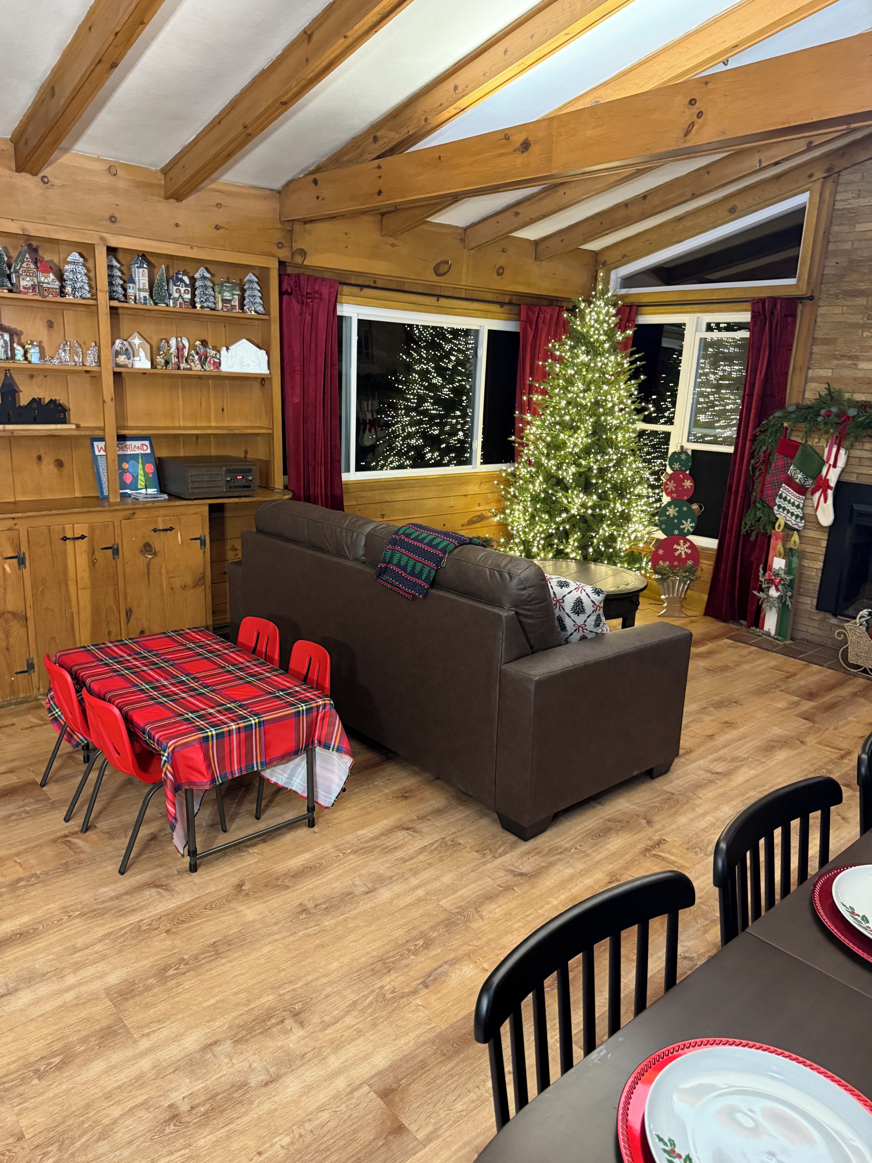A cozy living room with a wooden ceiling, a sofa, a dining table with chairs, and a Christmas tree.