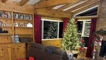 A cozy living room with a wooden ceiling, a sofa, a dining table with chairs, and a Christmas tree.