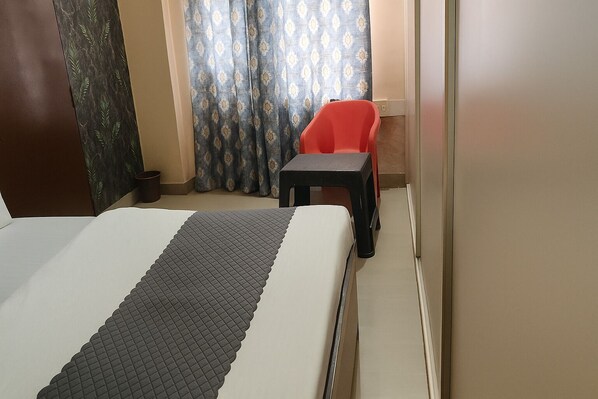 A hotel room with a bed, a small table, a red chair, and a curtain with a pattern. - Dev Hotel Mansarovar (Jaipur)