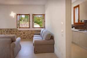 Ein Zimmer mit Steinwand, einer Couch, einer Küchenzeile und zwei Fenstern mit Blick auf Grünflächen.