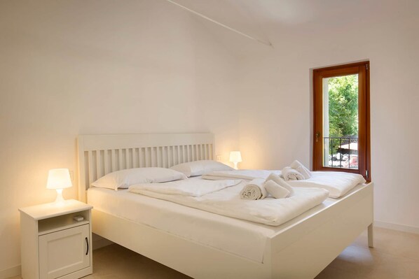 Ein ordentlich bezogenes Bett mit Kissen und Handtüchern, ein Nachttisch mit Lampe und ein Fenster mit Blick auf Grünflächen.