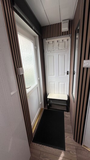 A narrow hallway with a door leading to a room, striped walls, a black mat at the entrance, and a small shelf with hangers. - Stylish Micro-Apartment with Secure Parking – Fast Train to London Central (Slough)
