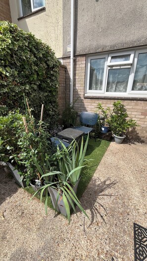 A garden area with a small table and chairs, potted plants, and a hedge. - Stylish Micro-Apartment with Secure Parking – Fast Train to London Central (Slough)