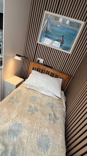 A bed with a floral bedspread, a bedside lamp, a power outlet, and a framed picture on the wall. - Stylish Micro-Apartment with Secure Parking – Fast Train to London Central (Slough)