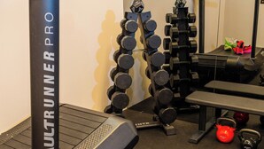 Ett gym med ett löpband av modellen AssaultRunner Pro, ett set hantlar, kettlebells och en hopfälld träningsmatta märkt med texten ”20 % CENTR”.