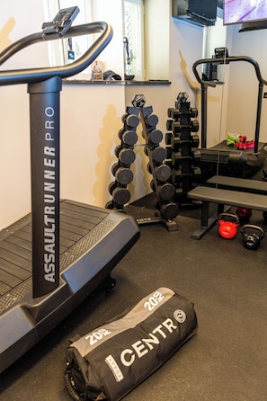 Ein Fitnessraum mit einem AssaultRunner Pro Laufband, einer Hantelbank, Kettlebells und einer zusammengefalteten Trainingsmatte mit der Aufschrift '20% CENTR'.