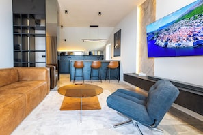 A modern living room with a leather sofa, a blue armchair, a glass coffee table, and a kitchen in the background.