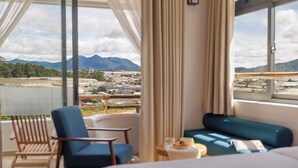 A room with a bed, a blue sofa, and a chair. There is a view of mountains and a cityscape through large windows.