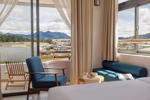 A room with a bed, a blue sofa, and a chair. There is a view of mountains and a cityscape through large windows.