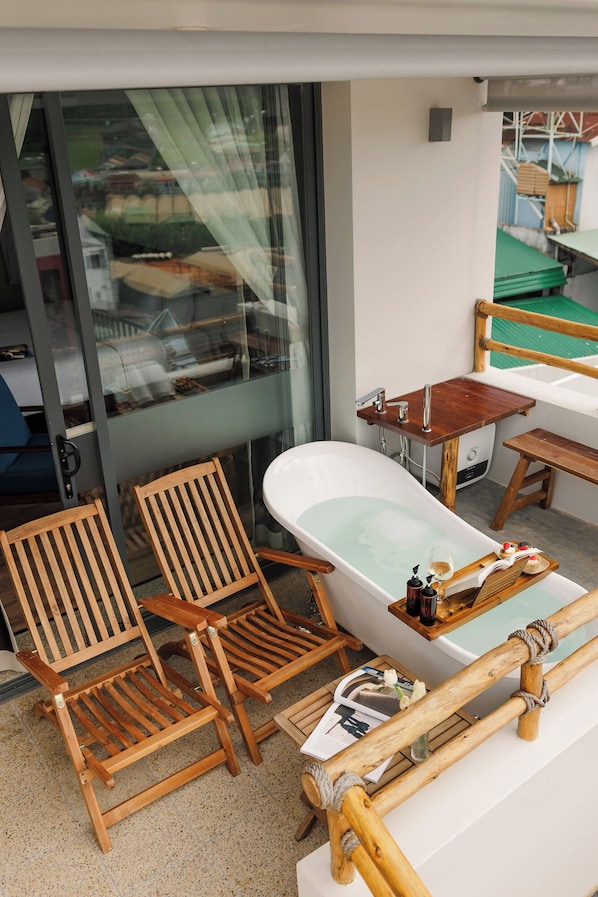 Un balcón con puerta de vidrio, sillas y mesa de madera, una bañera y un pequeño banco de madera.