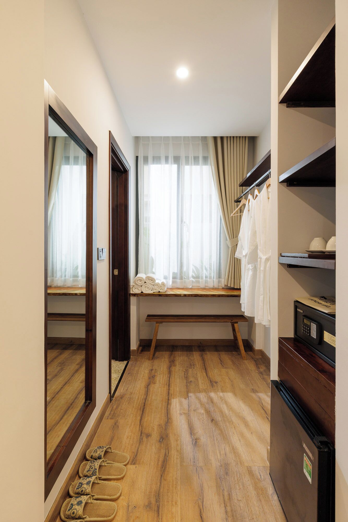 Un couloir avec un plancher en bois, un banc, une fenêtre avec des rideaux, un miroir et un porte-chaussures.
