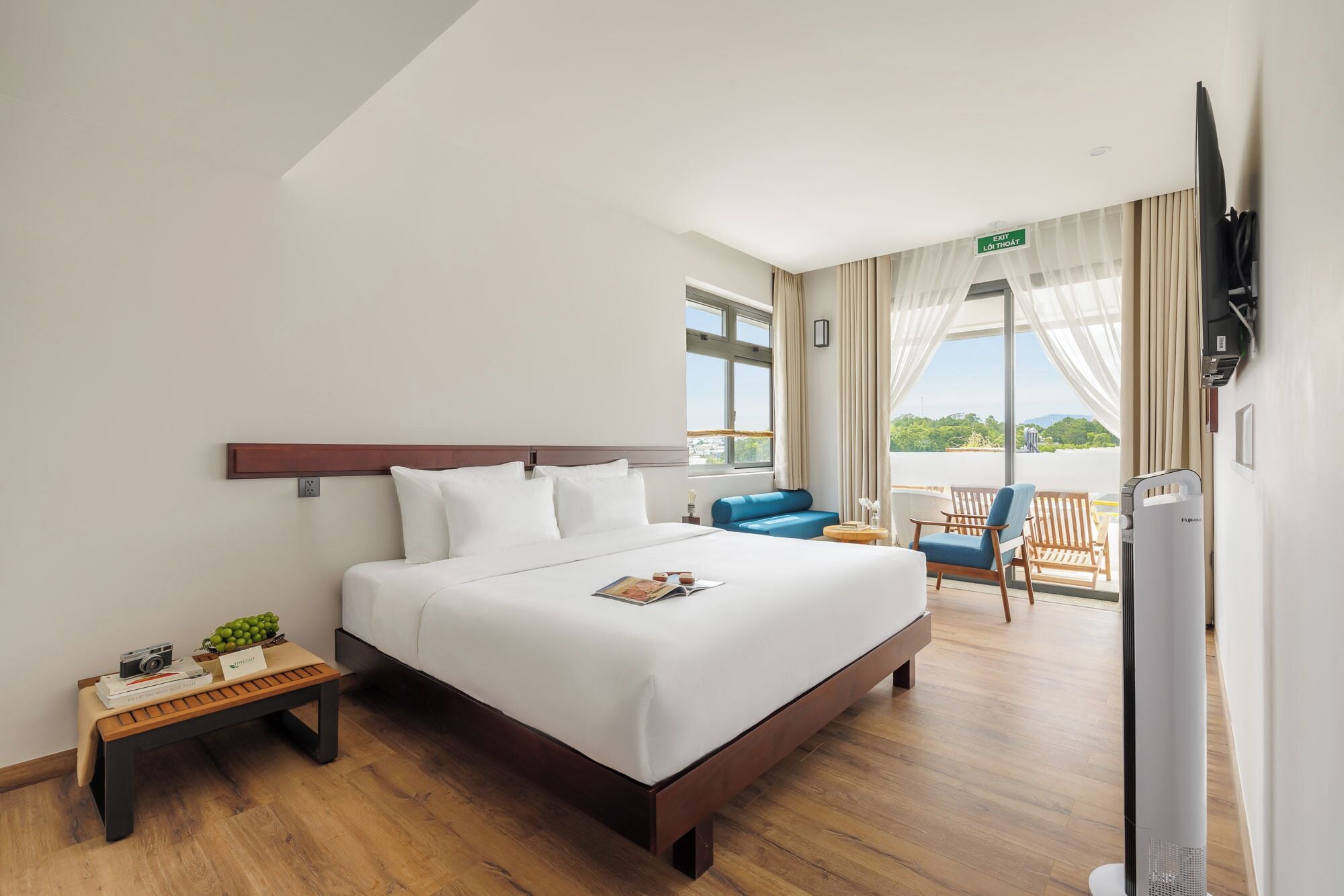 Habitación de hotel con cama, una mesita de noche con lámpara, una silla azul, balcón con vistas y televisor.