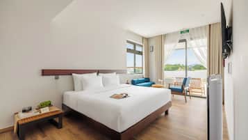 Habitación de hotel con cama, una mesita de noche con lámpara, una silla azul, balcón con vistas y televisor.