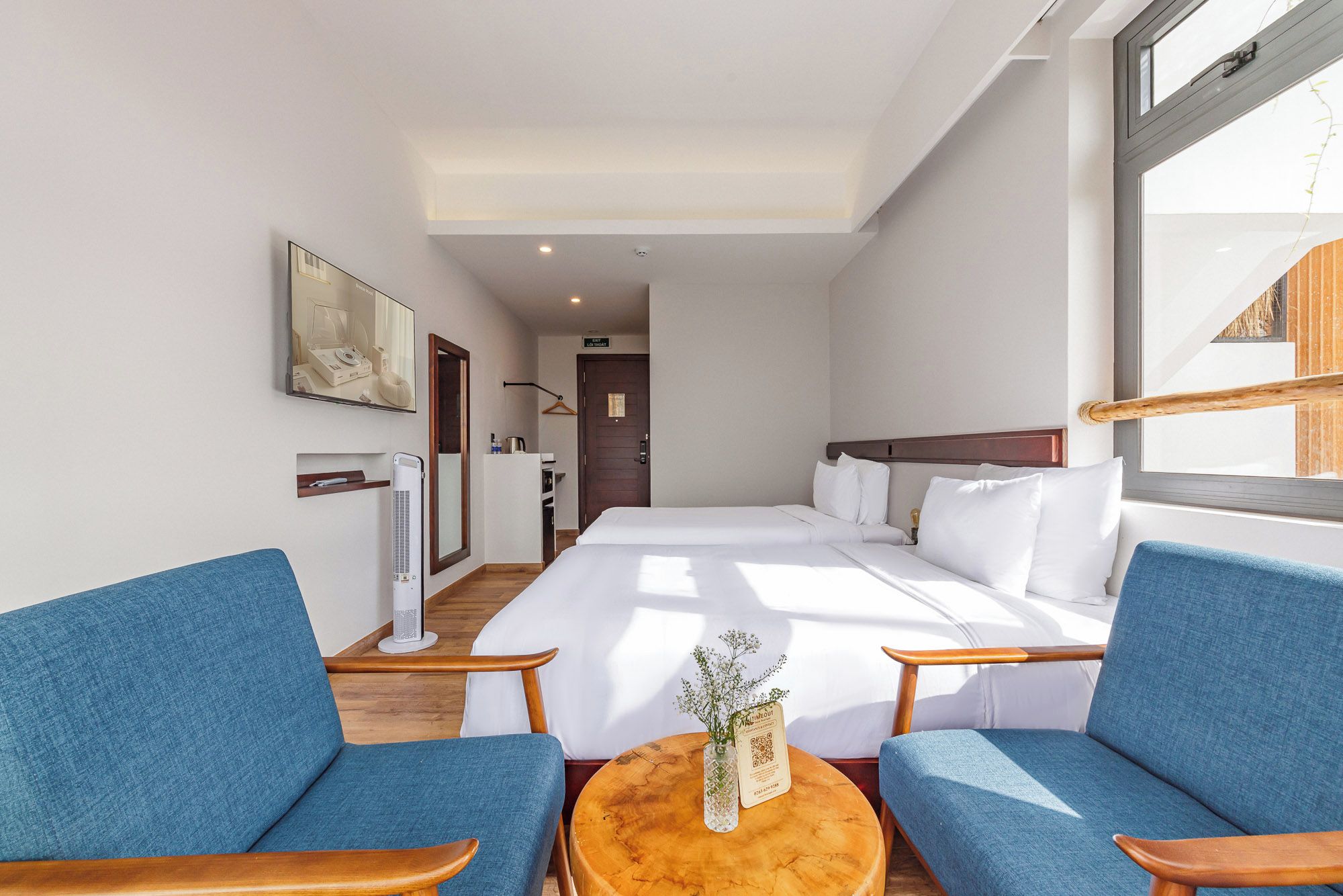 Habitación de hotel con cama, dos sillones azules, una mesita redonda con un florero y una ventana.