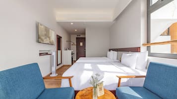 Habitación de hotel con cama, dos sillones azules, una mesita redonda con un florero y una ventana.