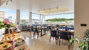 A spacious dining area with multiple tables and chairs, large windows offering a view of the cityscape, and a buffet table with various food items.