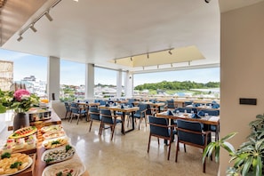 A spacious dining area with multiple tables and chairs, large windows offering a view of the cityscape, and a buffet table with various food items.