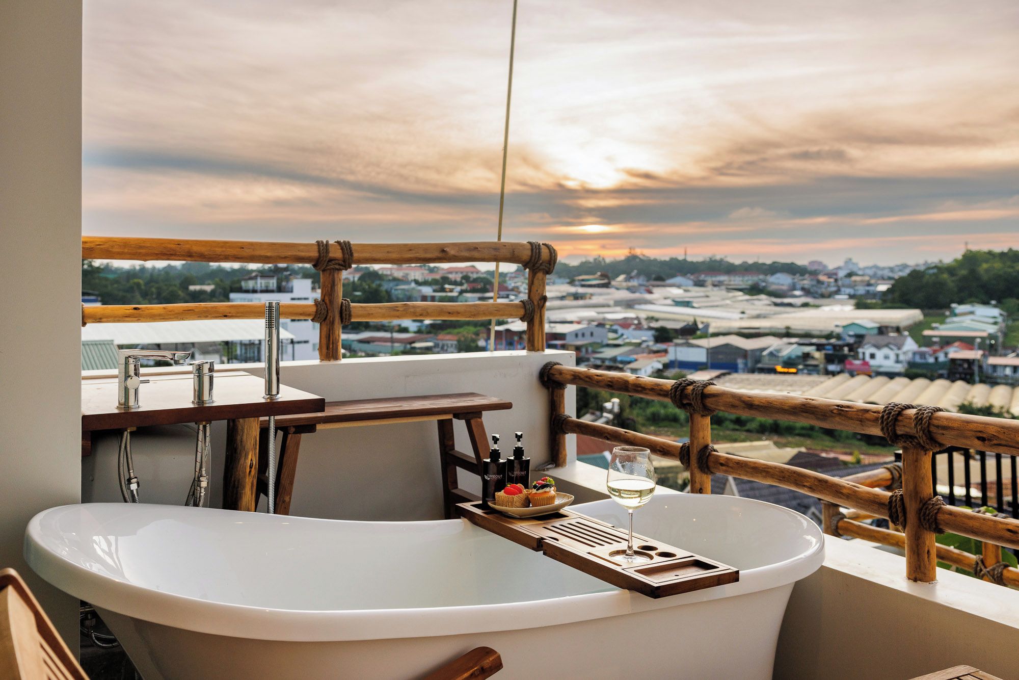A bathroom with a bathtub, a sink, and a wooden railing overlooking a cityscape at sunset.