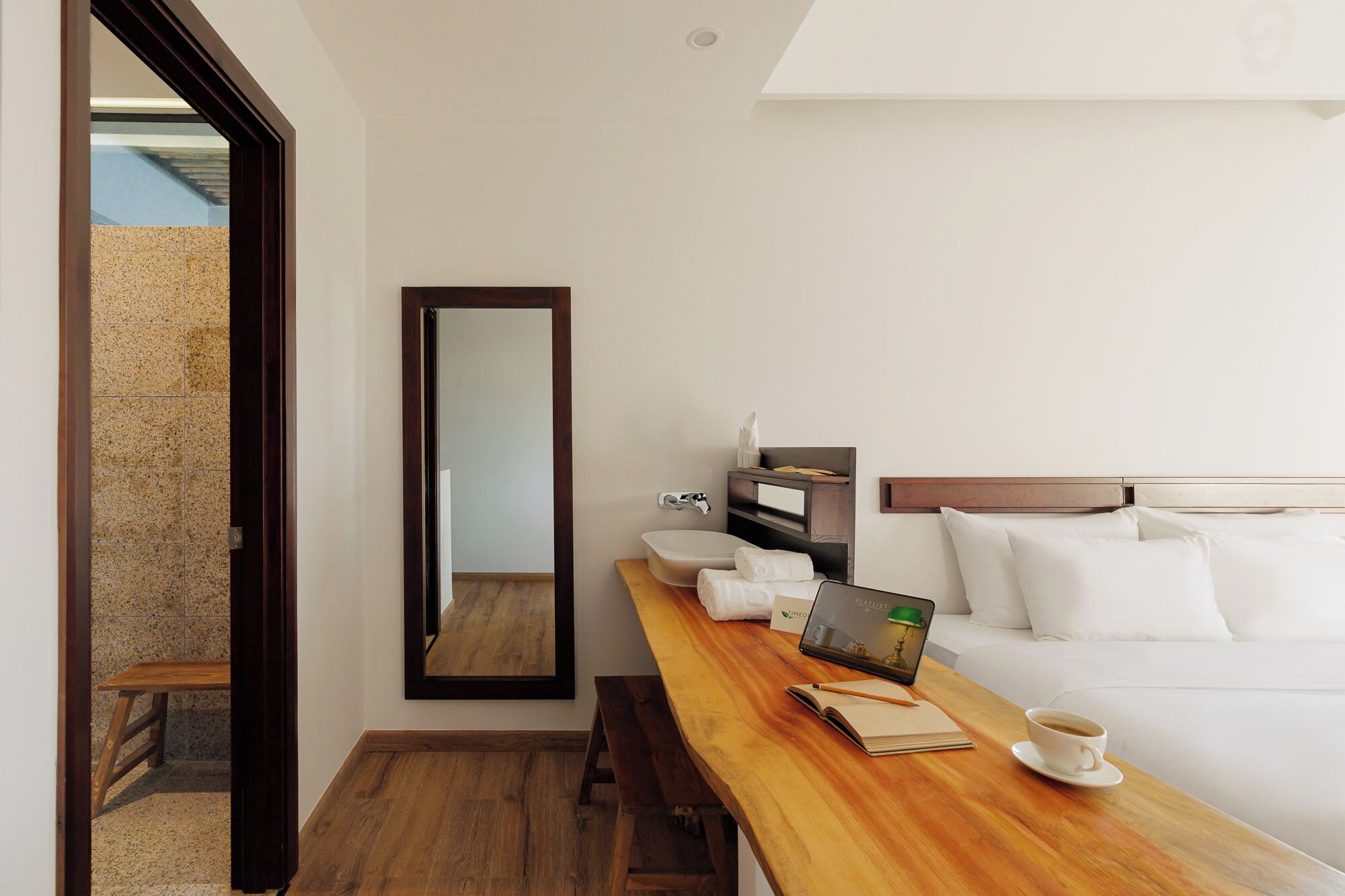 Une chambre d’hôtel avec un bureau en bois, un ordinateur portable, une tasse de café, un lit avec une literie blanche et un grand miroir.